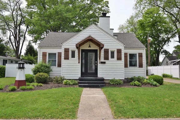 White cottage with brown shutters, black front door, and chimney surrounded by mature trees and landscaped lawn