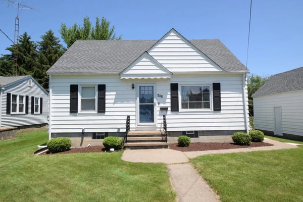 White single-story house with gray roof, black shutters, front porch, and manicured lawn with shrubs