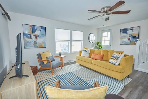 Bright living room with yellow sofa, mid-century armchair, striped rug, ceiling fan, and large windows with natural light