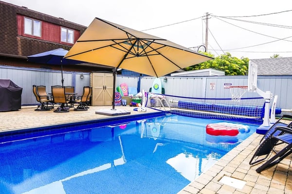 Backyard swimming pool with yellow umbrella, lounge chairs, basketball hoop, and inflatable toys