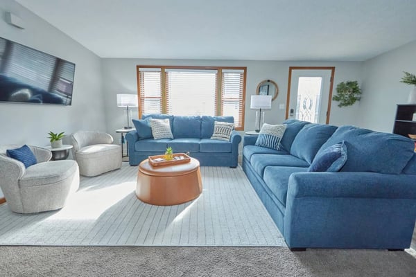 Bright living room with blue sofas, cream armchairs, round terracotta ottoman, and large windows with wooden frames