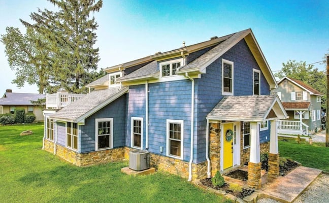 Blue cottage with yellow trim, stone foundation, and yellow front door on grassy lot with mature trees